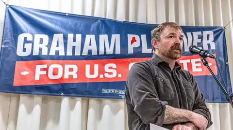 Graham Platner stands in front of a sign for his candidacy 