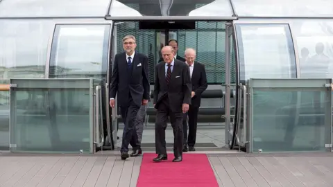 Getty Images Prince Phillip and other men in suits walking out of the glass i360 pod on to a red carpet.