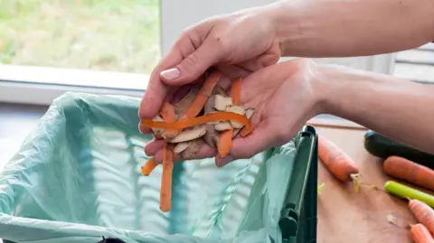A close-up of a person's hands holding carrot and potato peelings above a green food waste bag. More carrots can be seen on a chopping board.