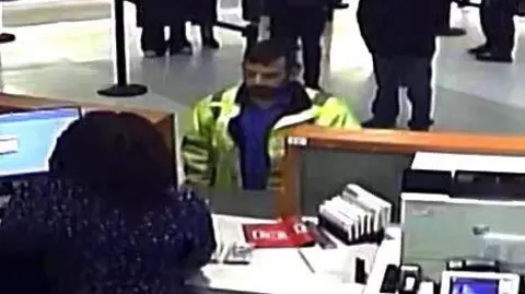 US Department of Justice A CCTV screenshot of Matthew McDonagh standing at a cashier's desk, wearing a yellow hi-vis jacket and a blue shirt. He has short black hair and a beard. We can see the back of the cashier's head, while others wait in line behind the man.