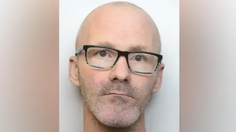 Gavin Collins who has a brown eyes, a bald head and grey stubble on his neck, chin and top lip and is wearing glasses with black frames  