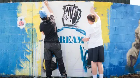Charlie Kirkpatrick/Supplied Two figures work on a blue spray-painted brick wall, filling in the outline of the back of a football player's shirt.