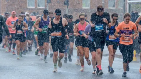 Tony Marsh Marathon runners in Edinburgh fight through the heavy rain. They are dressed in running gear and travelling along a flat road.
