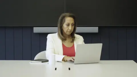 Moni Mannings, a woman with short brown hair wearing a cream jacket and coral top, sat at a white desk working on a silver laptop