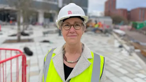 Professor Judith Squires is a middle-aged woman with white hair and green glasses. She has a chunky necklace on and is wearing a hi-vis jacket with a branded University of Bristol hard hat. She is smiling.