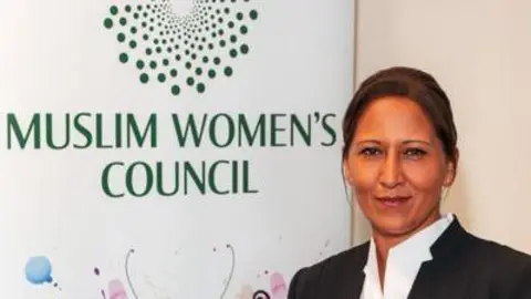 Bana Gora, a South Asian woman with brown hair. She is wearing a black blazer and stood in front of a sign which reads Muslim Women's Council.