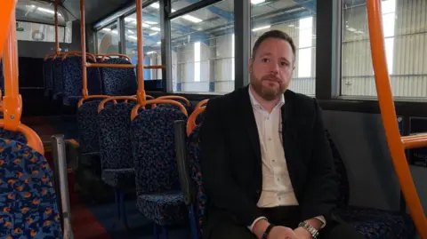 Tom Waterhouse is sitting on a bus in a storage shed. He is wearing a dark-coloured suit and white shirt.