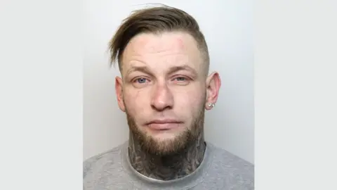 Wiltshire Police A police mugshot of Casey Delaney. He is wearing a grey t-shirt and standing against a grey background. His brown hair swept to the right on top with the sides shaved. He has blue eyes, a brown trimmed beard with no moustache and two diamond earrings in his left earlobe. He is looking at the camera with a blank expression.