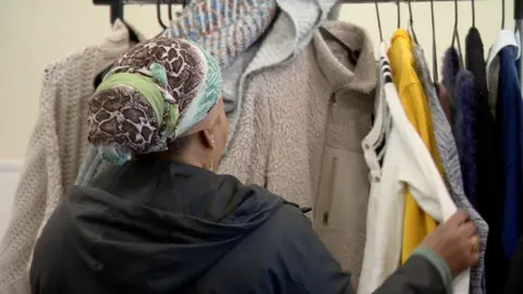 A woman wearing a snakeskin brown, white, green and blue scarf tying up her hair browses a rail of coats with her back to the camera
