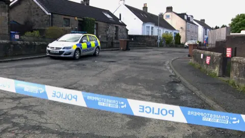 Police car parked outside a bungalow in the middle of a street. There is blue and white tape across the road saying POLICE and giving the Crimestoppers phone number. 