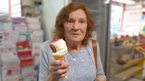 Eileen is holding an ice-cream cone in her hand and looking at the camera. She has brown hair and is wearing a grey top.