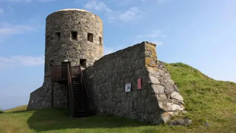 BBC Tower with wooden stair leading up to the opening of the structure. A plaque on the side of the wall displaying facts. Built on a mound of grass. 