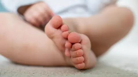 A close-up shot of a baby's feet which are sitting in a crossed legged position. It's hand can be seen in the background but is blurred. The Caucasian baby is wearing white clothing. 