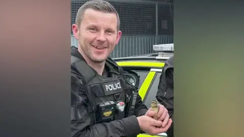 Cleveland Police PC Clough in his police uniform including a thick black stab vest smiles as he holds a small yellow duckling. 