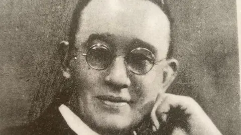 Holy Trinity Church An old black-and-white portrait of a man in a priest's white collar posing with a hand on his chin. He is smiling and wearing round glasses and his dark hair is swept back from his face.