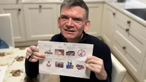 A man with greying hair and a black jumper on holds up a sheet with words and pictures on it. He also holds a pen in his hand. In the background you can see he is sitting in a kitchen 