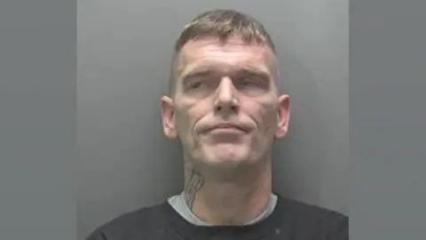 Cambridgeshire Police Andrew Jenkins in a custody photo. He is clean shaven and has cropped hair on his sides and longer light brown hair on top. He is wearing a black sweatshirt over a grey t-shirt and has a tattoo on the right side of his neck.