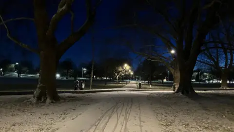 Camkingfisher/WEATHERWATCHERS Snowy paths lit up by street lamps. The two paths, which meet in the middle of the picture, are in a park or a green space. It is dark. There are three large trees at the point where the two paths meet. City lights can be seen in the distance.