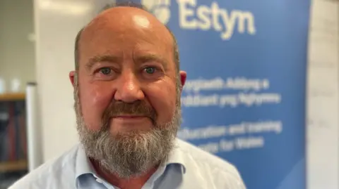 Owen Evans is looking into the camera and standing in front of an Estyn sign. He has a grey beard and is wearing a blue shirt.