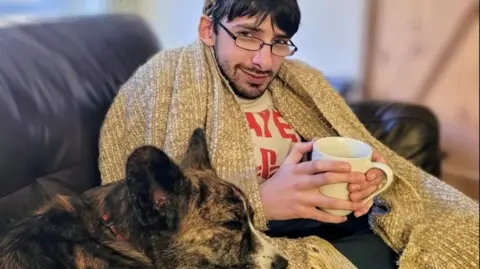 Family handout Daniel is sat on a settee with a blanket around him and a dog resting on his lap. He is holding a mug. He has glasses and a short beard.