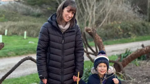 A woman in her thirties and her young son in a woodland area. The young boy is holding a stick and in the background there are tree branches and saplings covered by tree guards. 