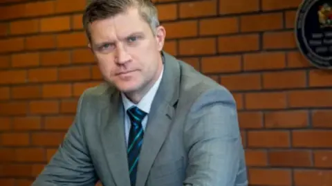Richard Cooke is wearing a grey suit, white shirt and blue and navy striped tie. He is seated, looking at the camera and leaning on a desk. 