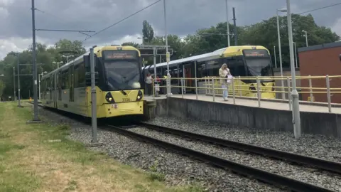 Two yellow Metrolink trams are stationary at platforms.