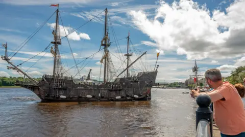 Derry and Strabane Council A man stands on a quayside looking at The Galeón Andalucía, a replica of a Spanish 18th century boat as it moors on a quay. It is a tall ship type vessel with high masts and big sails