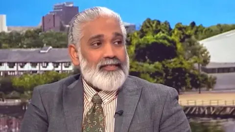 Dr Sakthi Karunanithi in a TV studio, in front of a screen showing rows of huts and some trees under a cloudless blue sky. He is facing right and has long, neat white hair tied back and a beard, and is wearing a gray suit, a white and brown striped shirt, and a green tie with flowers.