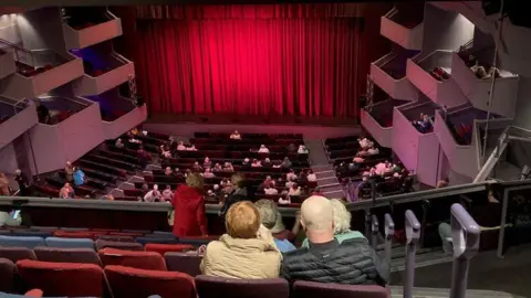 Martin Heath/BBC The Derngate auditorium with a red curtain across the stage and people sitting in the stalls and circle. There are pink boxes on either side protruding over the stalls. There is a spotlight on the curtain.