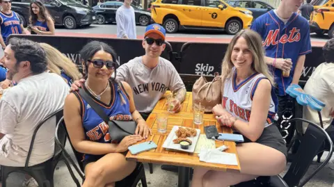 Grace Eliza Goodwin/BBC a group of three people sitting at a bar patio, wearing Knicks gear