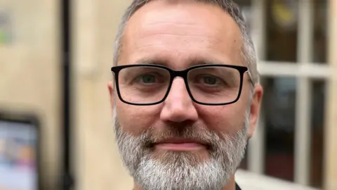 Simon Allen with a grey beard and black rimmed glasses smiling at the camera