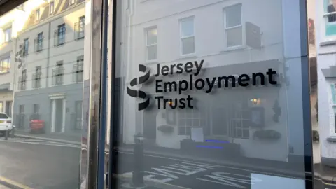 A window displaying a Jersey Employment Trust logo. Buildings opposite the window are in the reflection. 