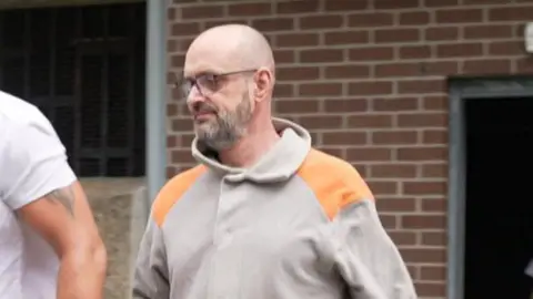 Paul Lewis pictured leaving Merthyr Crown court. He wears a grey and orange hoodie. He is bald with a grey beard and wears square reading glasses. Paul Lewis was alleged to have paid Dominique Saunders to arrange the murder of his wife Joanne Atkinson. Both men denied the charges.