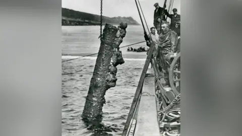 Museum of Cornish Life A cannon is hung on a chain partially out of the water at the side of a ship as divers watch on.