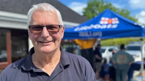 A man stands in front of a small marquee. He's in a navy polo shirt and is wearing sunglasses. He's smiling. Behind him the Blue branding of the RNLI can be seen on the marquee, as people stand under it in shade from the bright sun. 