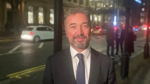 Chris smiling at the camera. He is wearing a black suit jacket, white shirt and blue tie. He is standing on the pavement, with the street behind him. On the street, there are cars and a white car is visible behind him.
