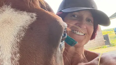 Sue, a smiling woman in a baseball cap, stands next to a horse. The horse is closer to the camera, with only its eye and forehead visible next to it.