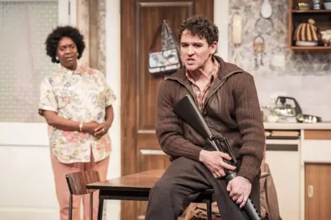 Marc Brenner Robbie O’Neil holding a rifle and perching on a table, speaking to Anita Reynolds in her kitchen in a scene from The Assassination of Margaret Thatcher at Liverpool Everyman
