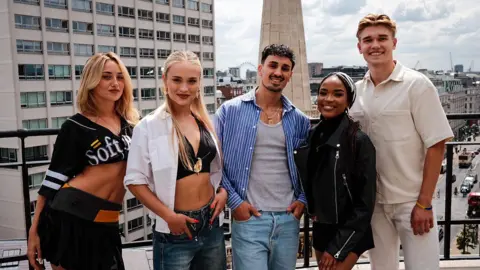 BBC/Ben McElligott Five dancers stand on a rooftop terrace overlooking a city centre. Office buildings, a street below and a landmark column are visible in the background.
