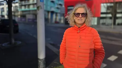 A woman with short blonde hair. She is wearing sunglasses and an orange coat. Behind her is a road and a big red building and a block of flats.