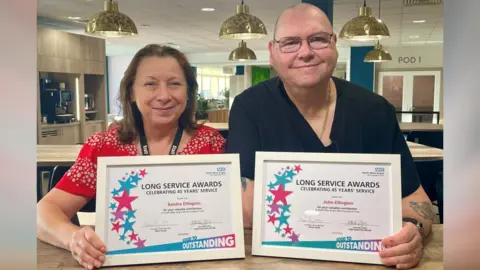 NWAFT Sandy Ellington, wearing a red blouse with a white dot pattern, and John Ellington, wearing a short-sleeved black top and gold necklace are pictured with a hospital cafeteria. Both are holding their white long-service certificates and smiling.