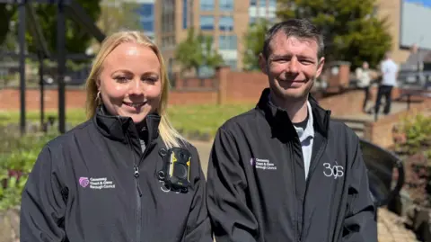 BBC A woman with blonde long hair and a man with short spiky brown hair smiling at camera. They are both wearing black jackets with logos of Causeway Coast and Glens Borough Council and 3GS. Sarah's jacket has a camera attached. There is a park in the background with people standing talking blurred in the background. 