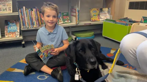 BBC Seven-year-old Alfie sits on the floor next to Rafa, the black Labrador. He is reading him a book called Party Games. A book shelf with other children's books on it is behind Alfie and Rafa.