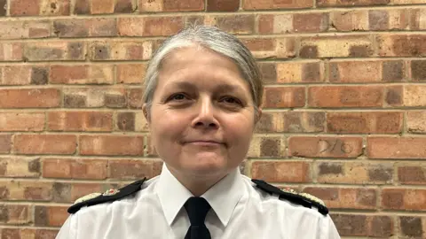 Sarah Crews photographed inside, against a brown brick wall. She is smiling slightly at the camera and has light grey hair pulled back away from her face. She is wearing a white shirt and a black tie, and has two large black shoulder pads on her shirt with the police emblem on it. 