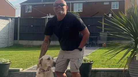 A man wearing sun glasses, a T-shirt and shorts, stands in a garden smiling with his right hand on a dog, which is sitting. Behind him is a lawn with a brown fence at the back. He is standing on a paved courtyard with flower pots on it and steps leading up to the lawn.