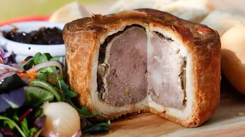 Getty Images/Bloomberg A photograph of a Melton Mowbray pork pie, with a slice missing, surrounded by salad to the left and a piece of bread to the right.