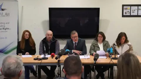 PA Media Kevin Winters with short greying hair and glasses wearing a dark grey suit flanked by three women and a man who are relatives of Katie Simpson with microphones in front of the table they're sitting at  