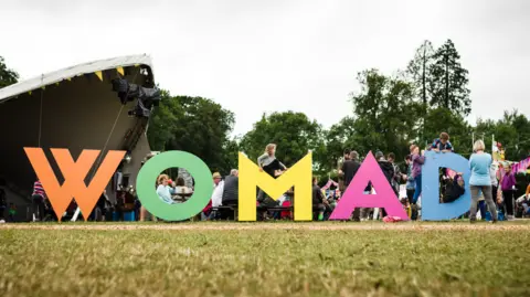 A field with a large music stage in the background and lots of people in front of it. In the foreground of the picture there are huge multi-coloured letters spelling out 'WOMAD' in capital letters