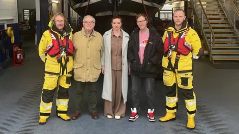 Six people stand in a lifeboat station in front of an RNLI lifeboat on its slipway. Two RNLI crew members in yellow protective gear stand at either end, with four visitors in everyday clothing between them. The lifeboat’s bow is raised behind them inside the boathouse.